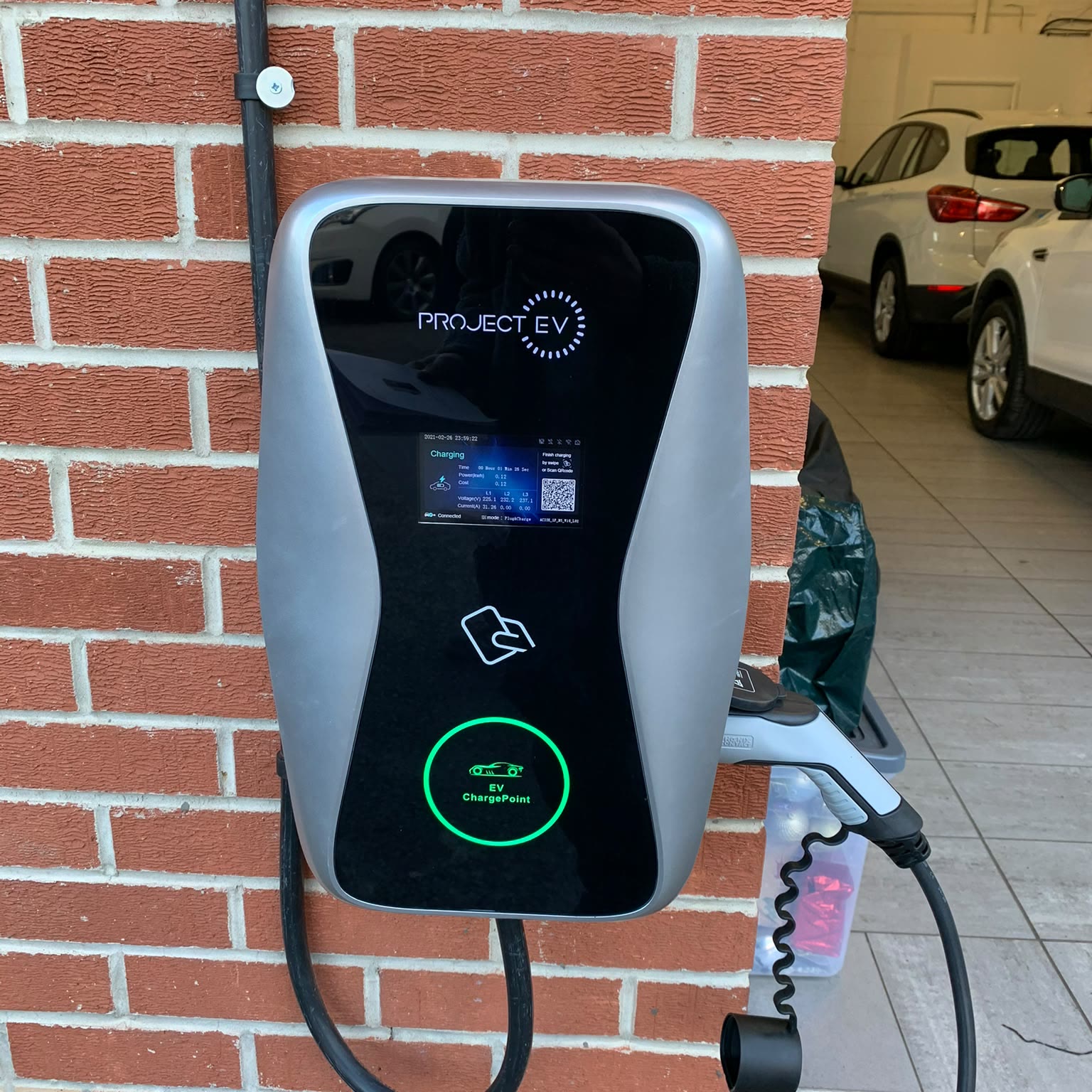 Wall-mounted electric car charger on a house wall, plugged in and charging, with the car on the drive behind