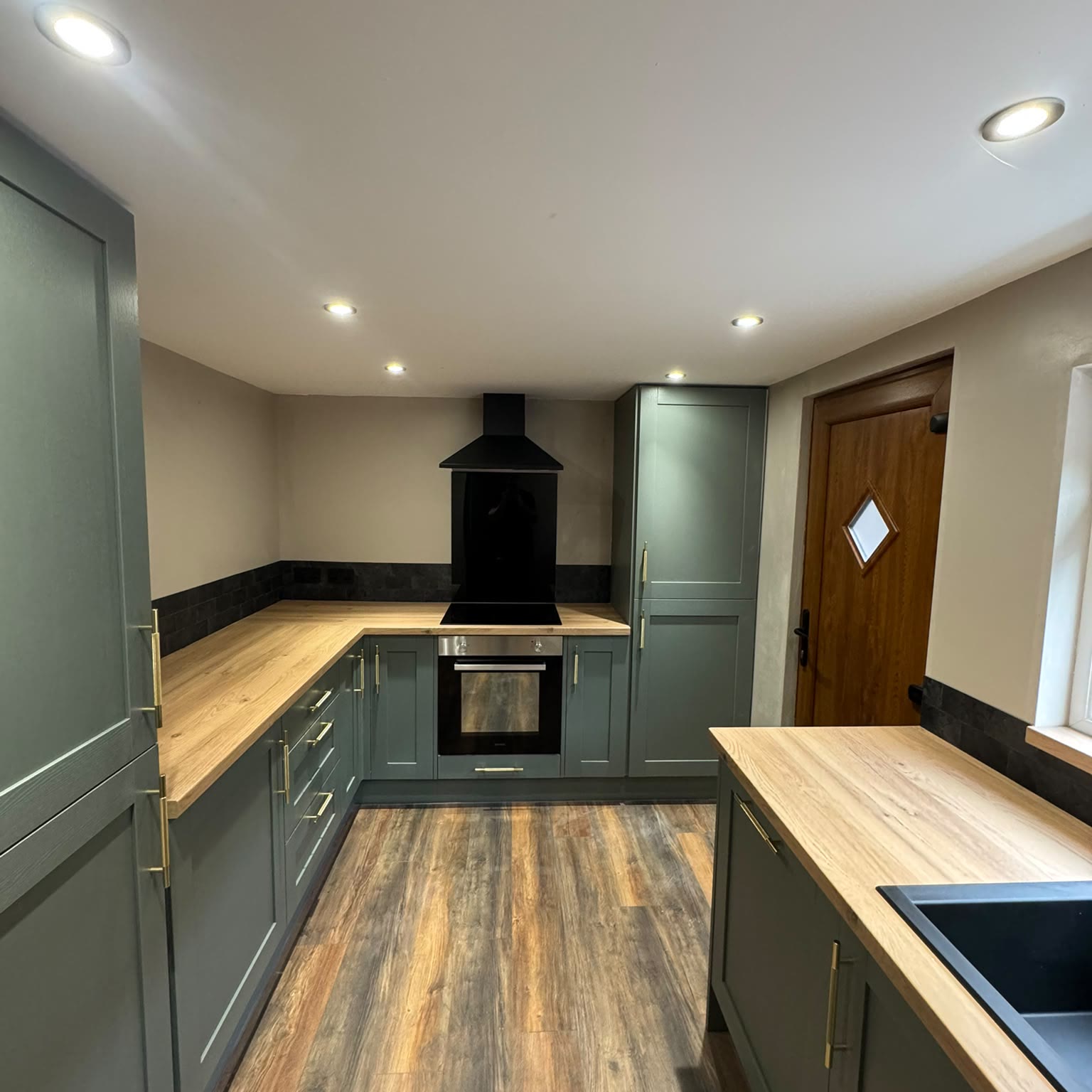 Finished kitchen in sage green with downlights through the ceiling, a wood-effect worktop and a lit extractor over the hob