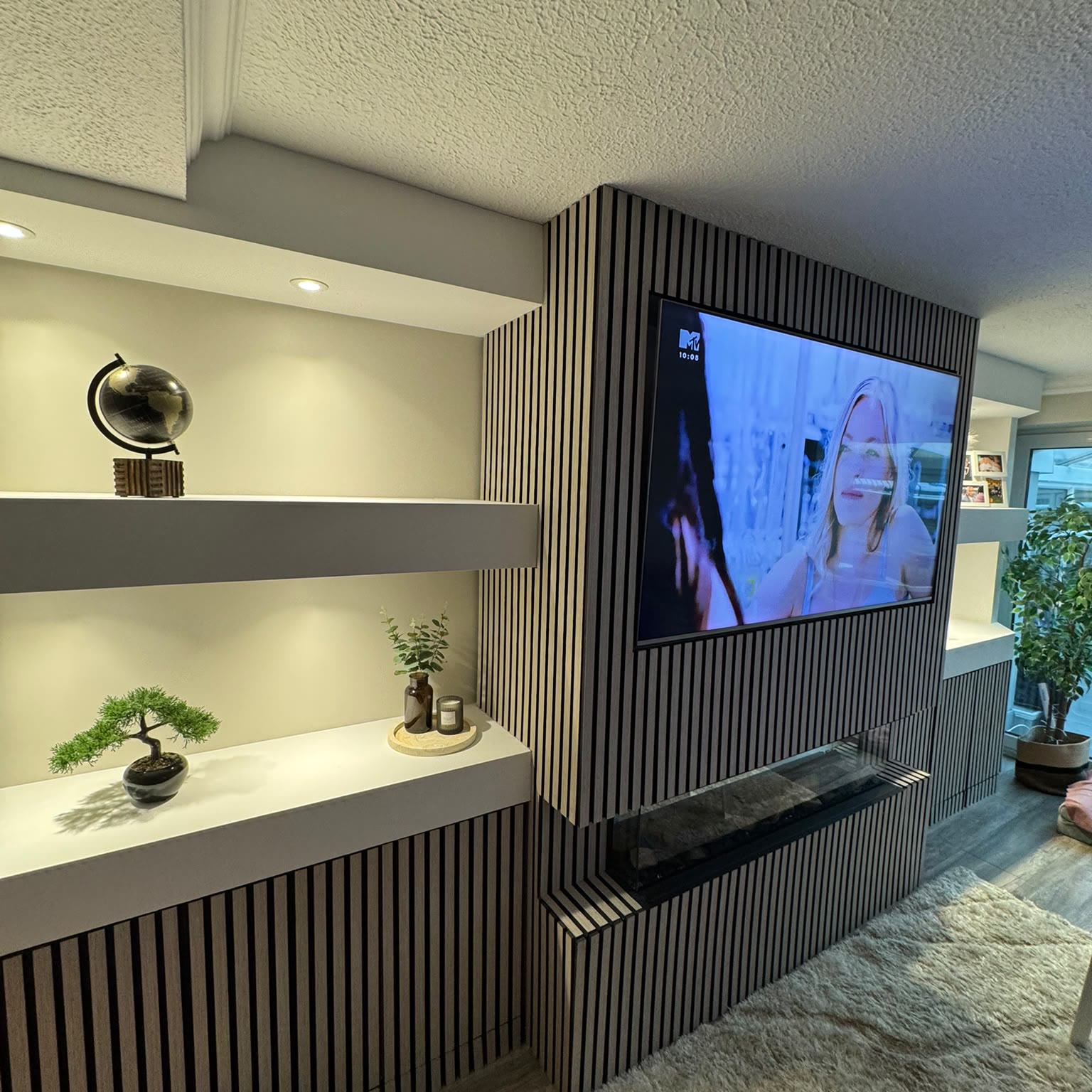 Living room media wall in slatted timber, with concealed lighting to the shelves, a wall-mounted television and an electric fire below