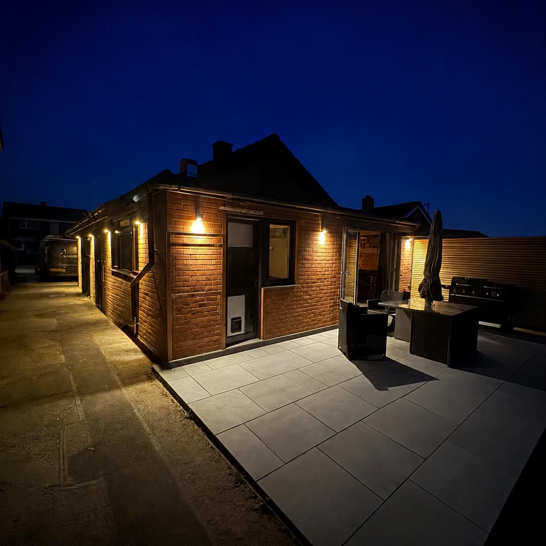 Garden room and patio lit at night by wall lights along the length of the building