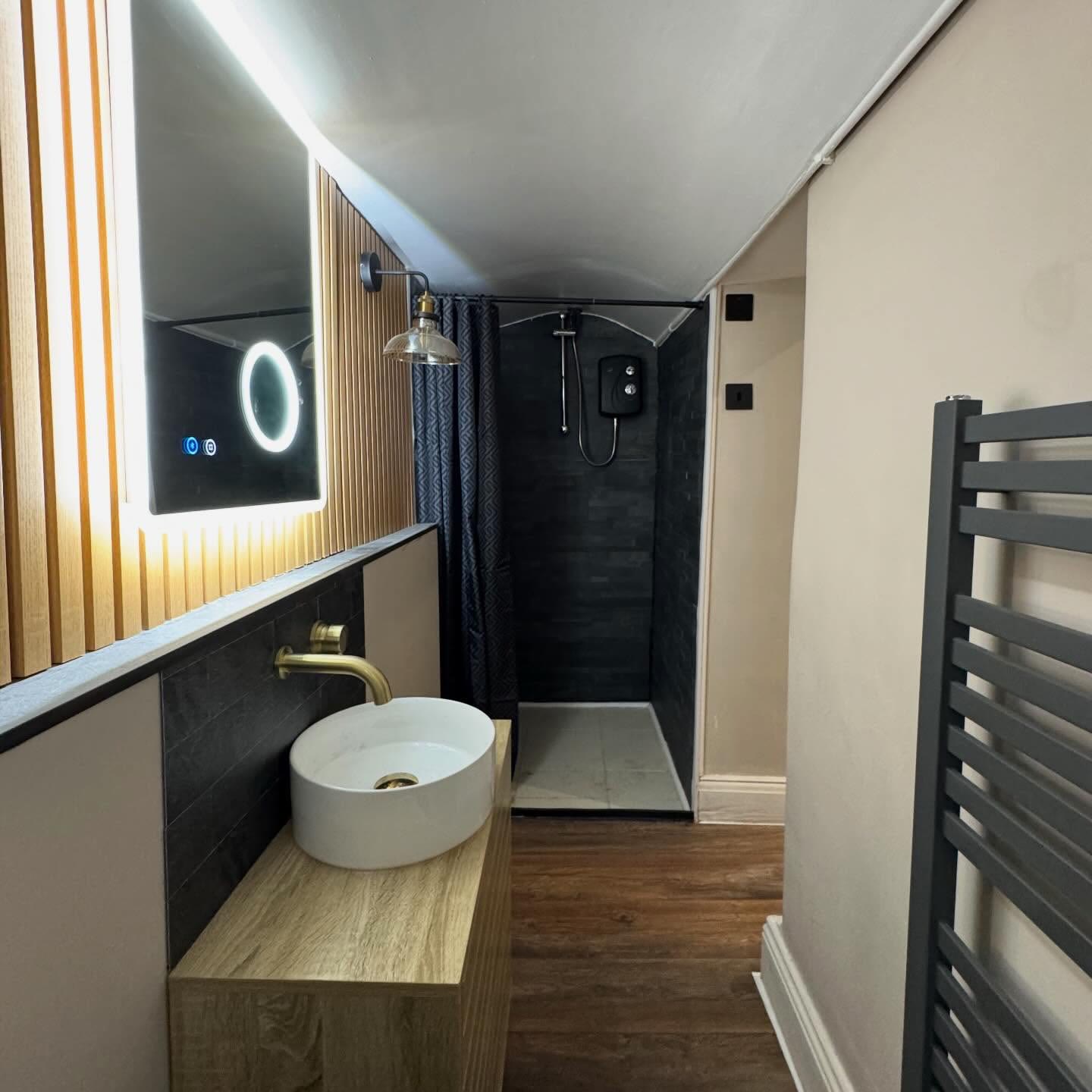 Bathroom with an illuminated LED mirror, brass wall light and an electric shower installed in a tiled recess